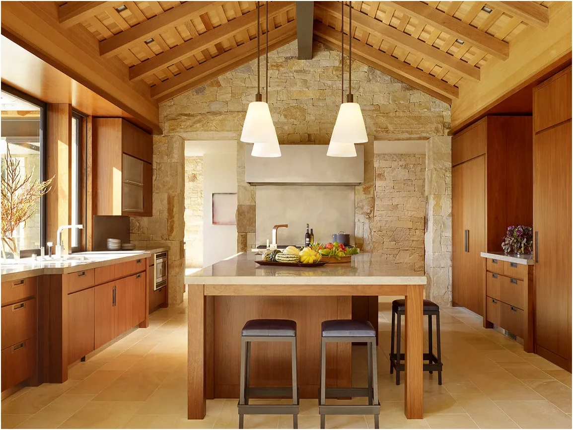 Honey-toned wood cabinets and ceiling framing surround a pale stone wall and floor, with a matching wood island and dark stools.