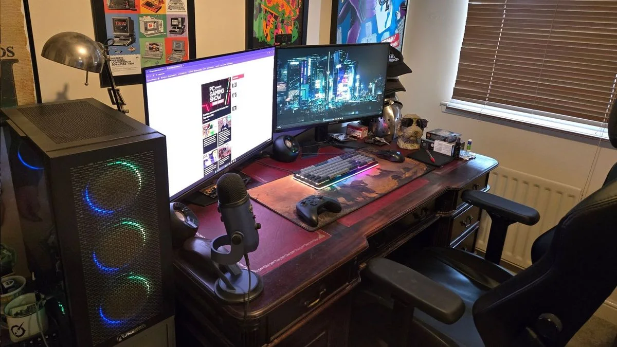 A dark wood desk with two monitors, a glowing black computer tower, a desk lamp, framed posters and a black chair.