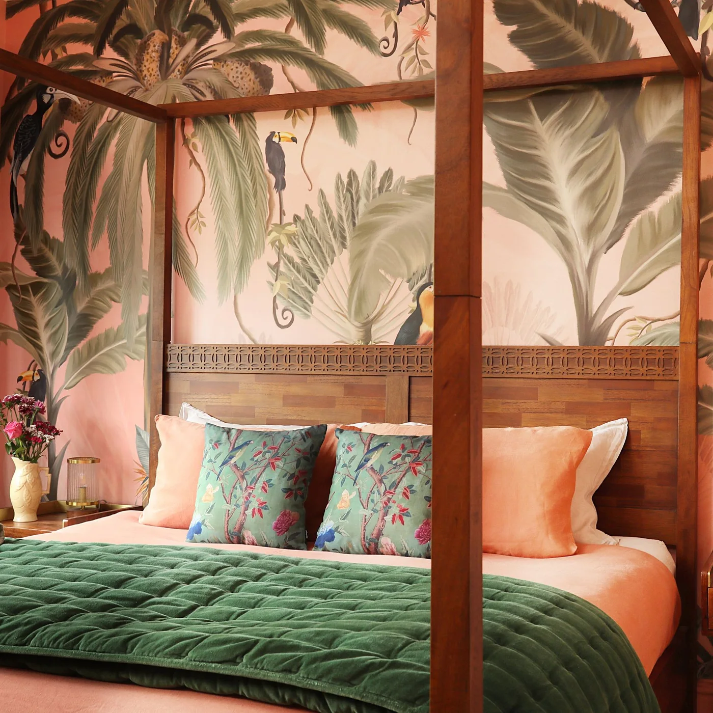 A wood four-poster bed with peach pillows and a green quilt stands against peach wallpaper filled with large green leaves and tropical birds