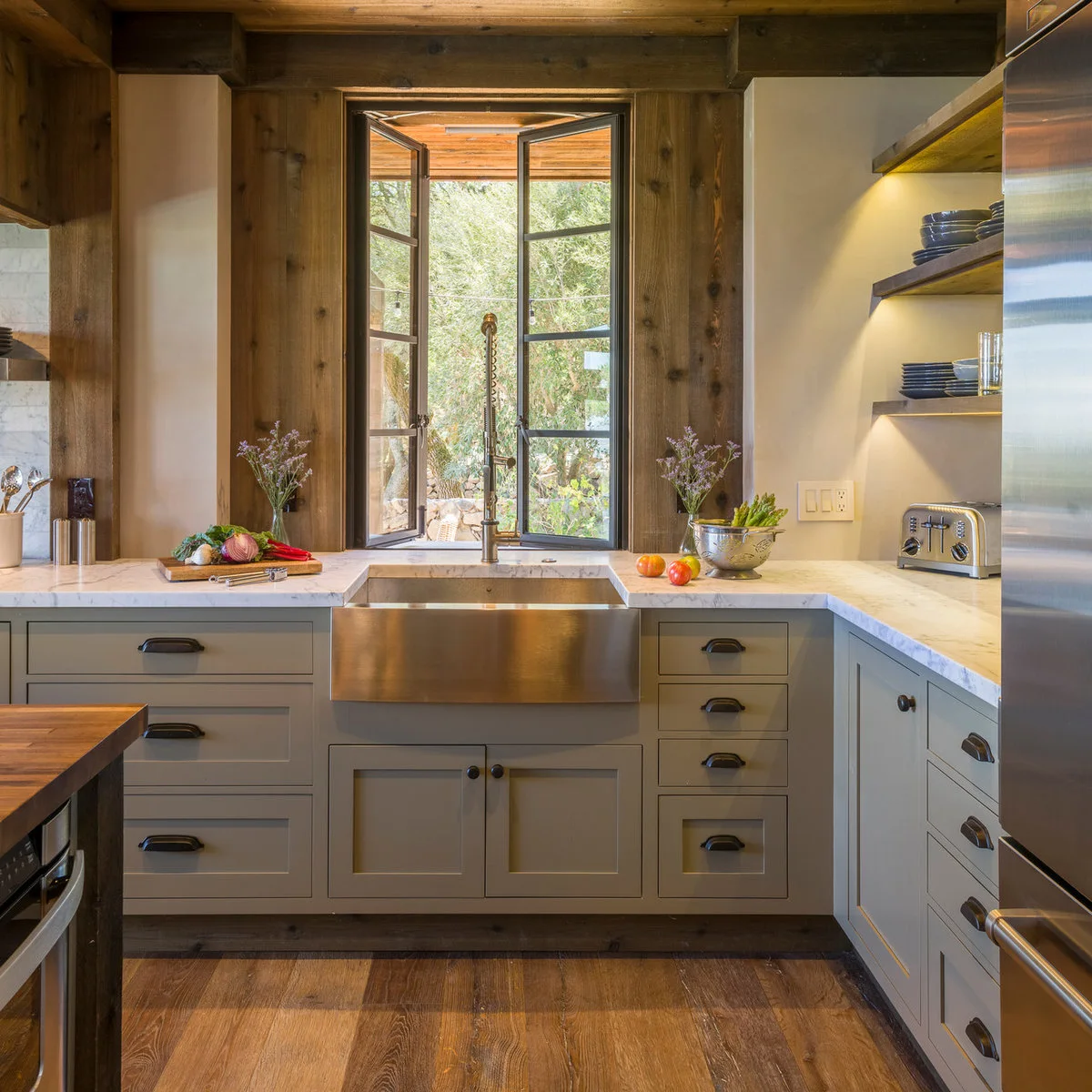 An inward-open pair of dark-framed windows above a metal apron-front sink and a tall spring faucet.