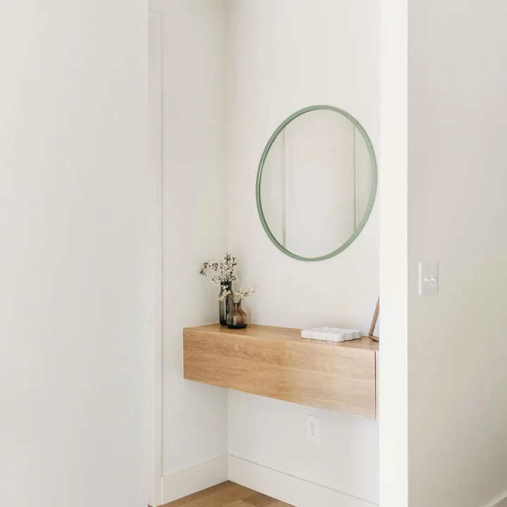 A floating wood console sits beneath a round pale green mirror in a narrow white hallway recess.