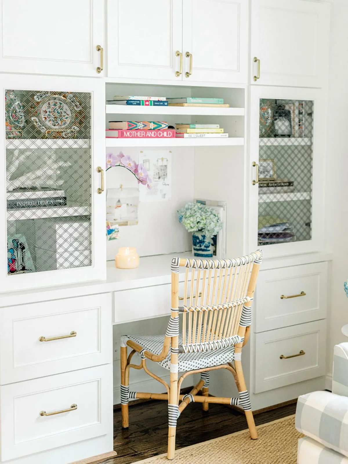 A white fitted desk is flanked by drawers and glass-front cupboards beneath upper cabinets, with a patterned woven chair and small task area.