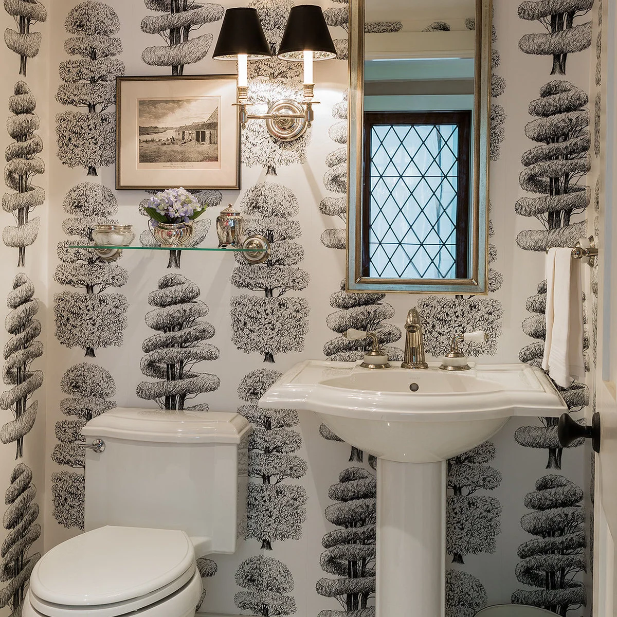 A white pedestal sink stands against black and white tree-patterned walls beside a short glass shelf and a narrow framed mirror.