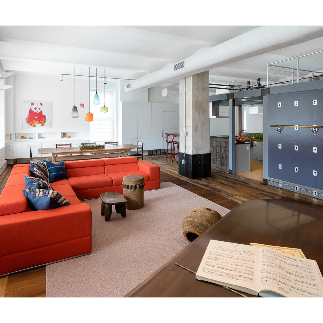 A bright orange sectional on a pale rug beneath exposed white ducts and a metal mezzanine