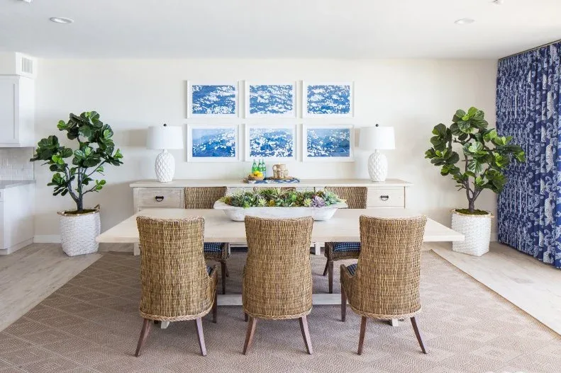 White dining room with woven chairs, blue artwork above a sideboard and matching lamps and plants.