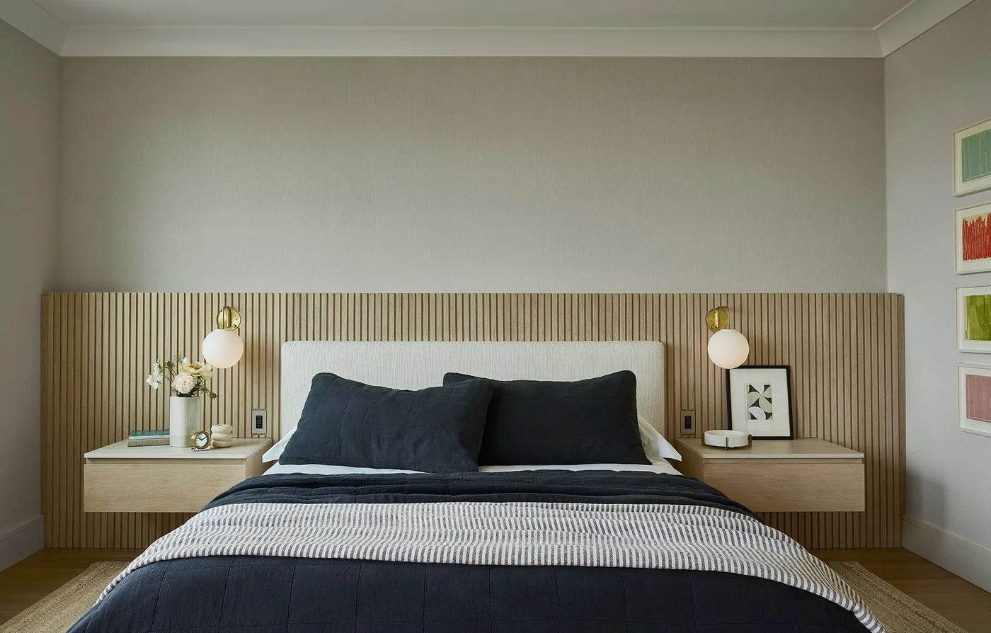 A pale bedroom has a wide wood-slat headboard wall, floating drawer tables, round white wall lights and dark blue bedding with a striped throw.