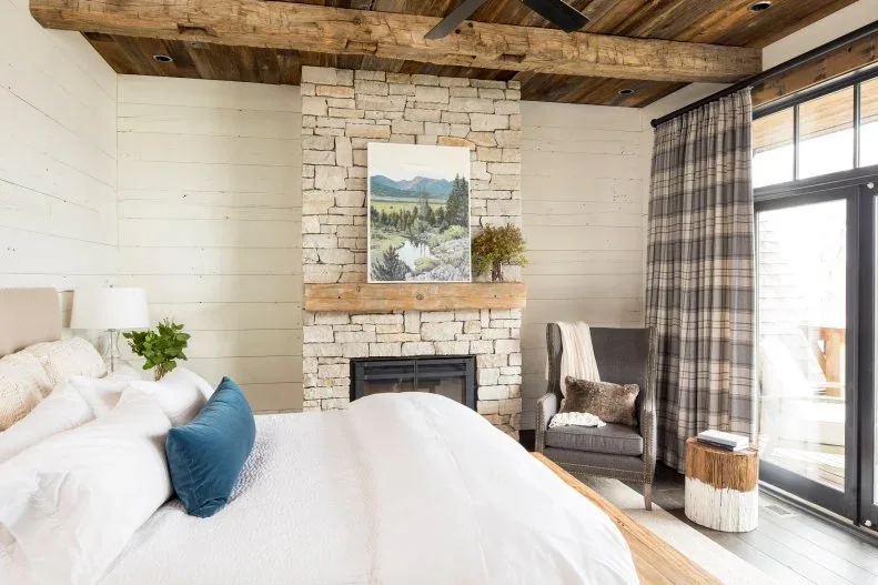 A bedroom stone fireplace wall and timber ceiling sit beside pale bedding, checked curtains and a gray chair.