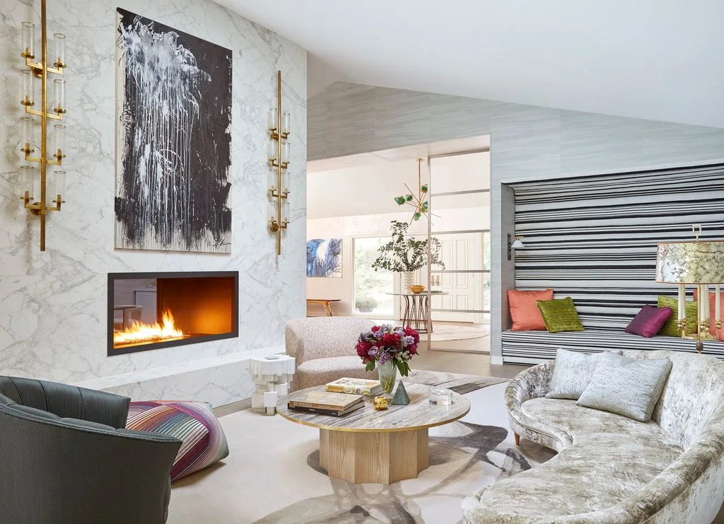 A rectangular fireplace opening sits within a pale veined wall beneath dark abstract artwork and slim gold-toned wall lights beside rounded pale seating