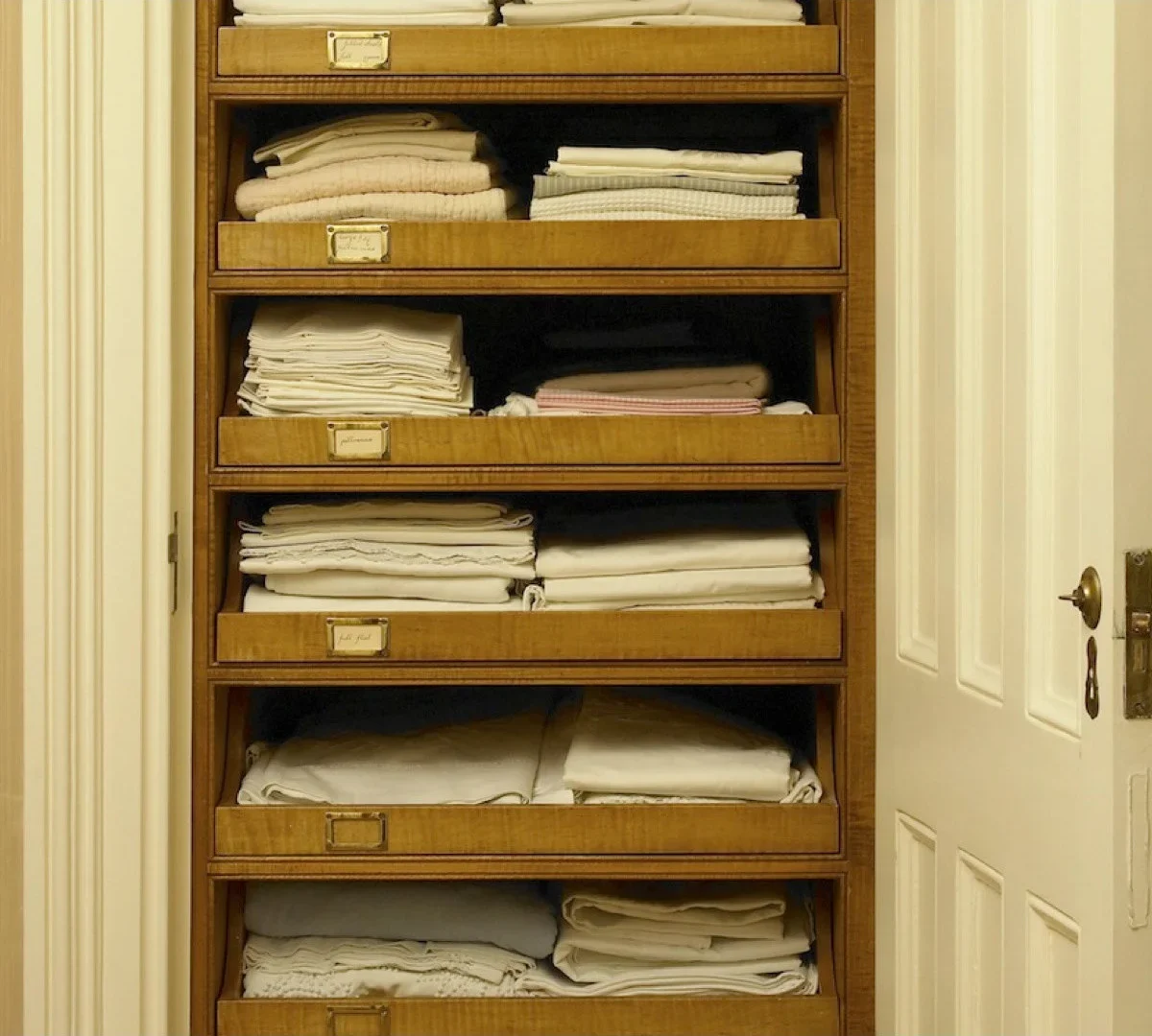 Wooden linen cupboard with shallow pullout trays holding folded sheets and towels