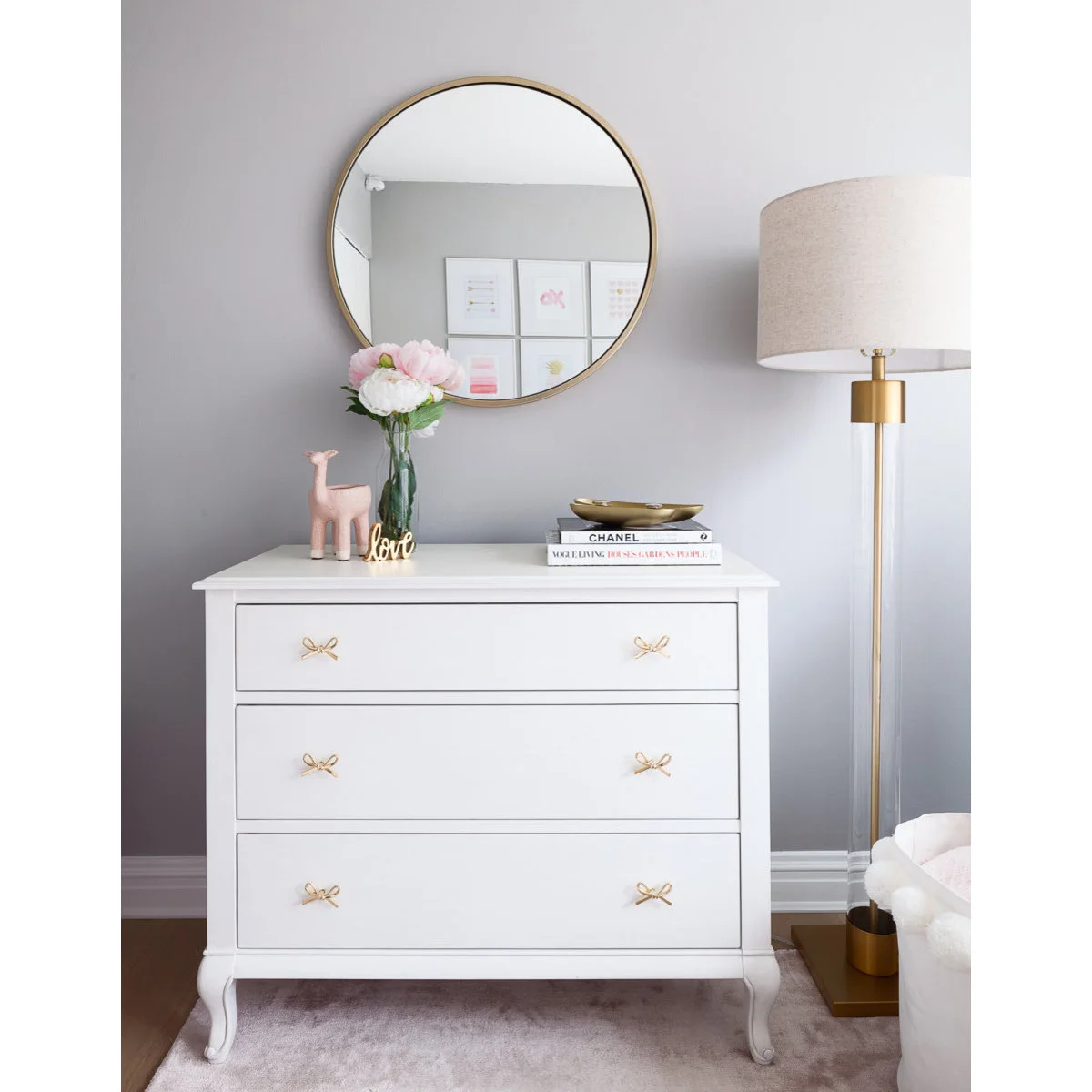 White nursery chest with gold-colored bow handles below a round mirror beside a floor lamp