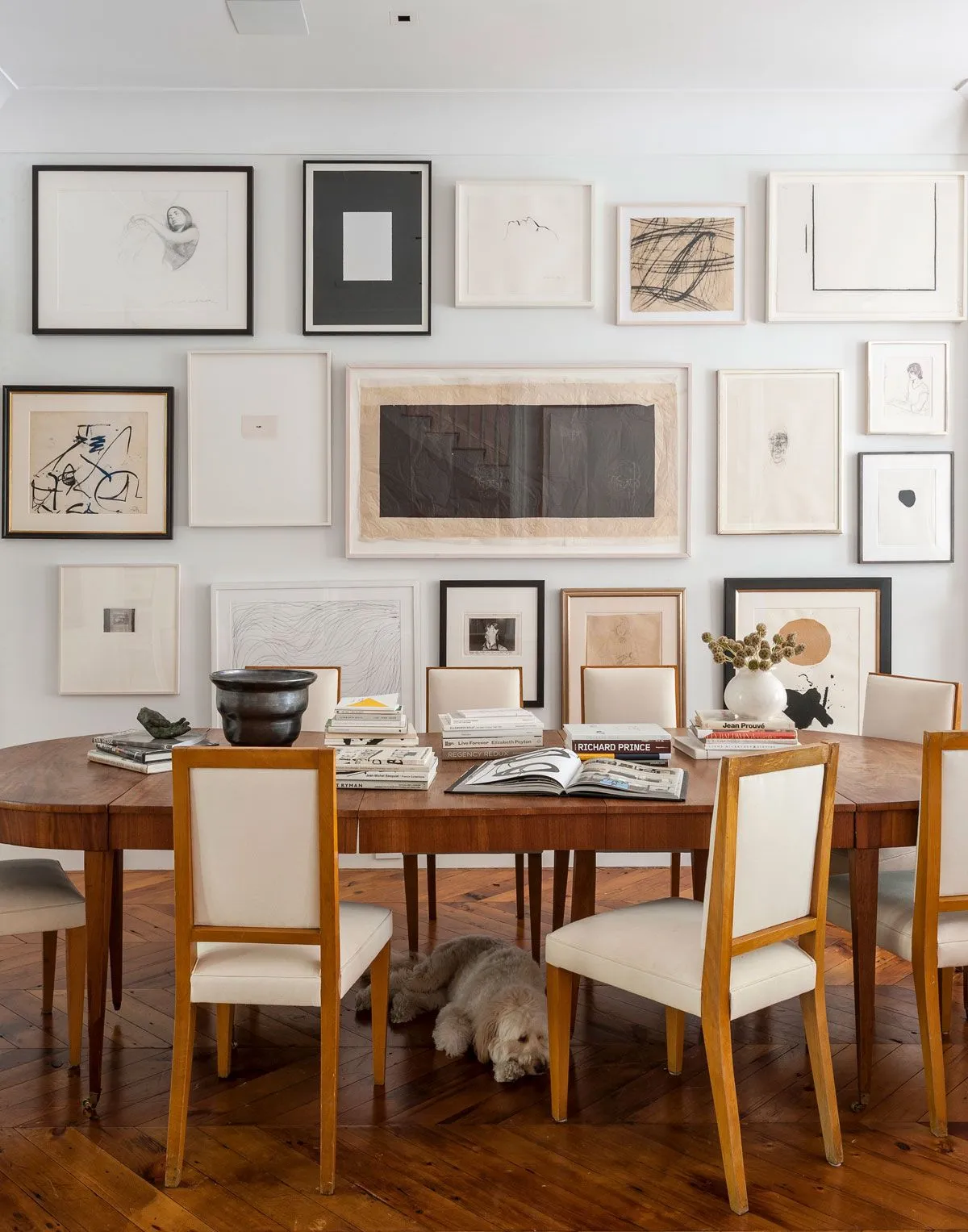 A timber dining table and pale upholstered chairs sit beneath a dense collection of framed pictures.