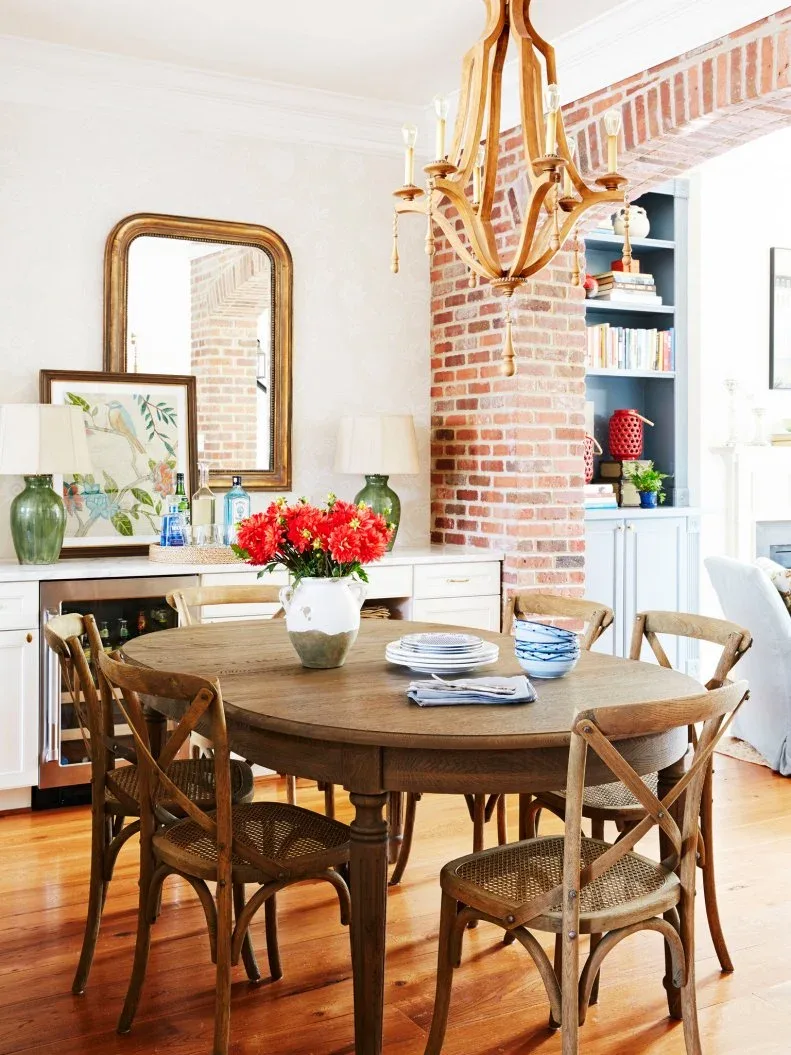 Pale wood-framed sideboard beneath a gilded mirror and green lamps beside a brick arch and round dining table
