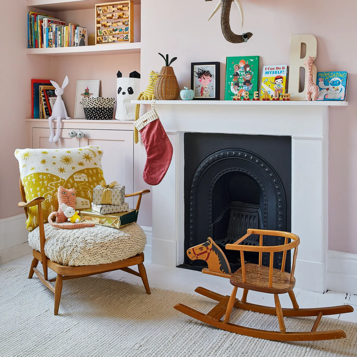 Pink nursery seating corner with yellow-patterned wooden armchair, pale fireplace, books and wooden rocking horse.