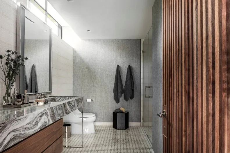 A bathroom with a veined counter and wood vanity has a tall wood-slat detail beside a glazed shower, gray wall and pale small-format floor tiles