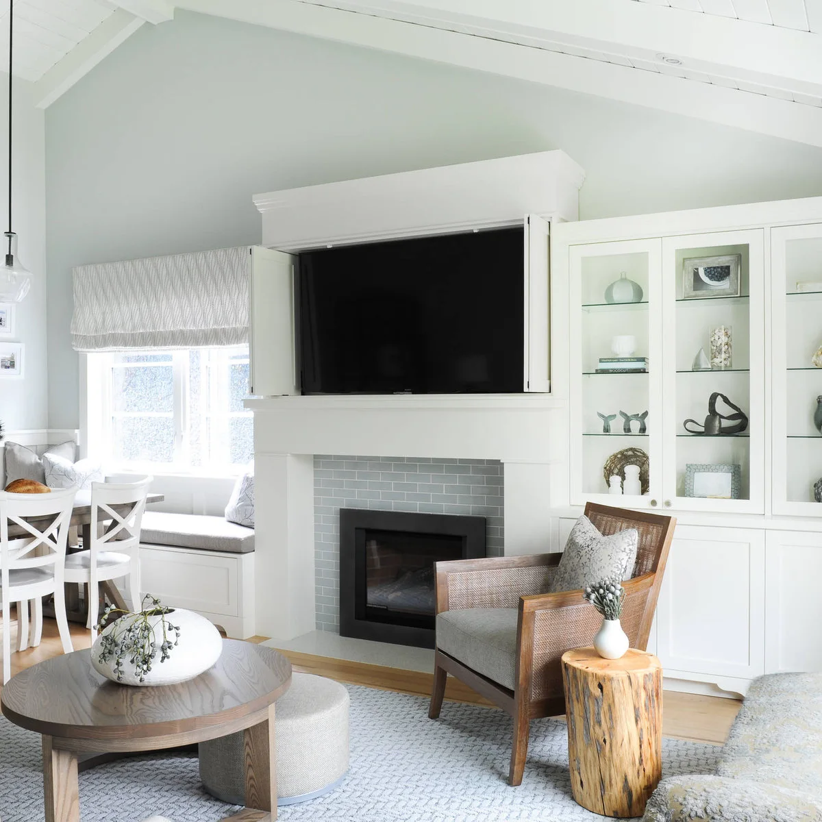 A pale room has white dining chairs beside a window bench, a large television in cabinetry above a fireplace and a woven armchair near a round coffee table.