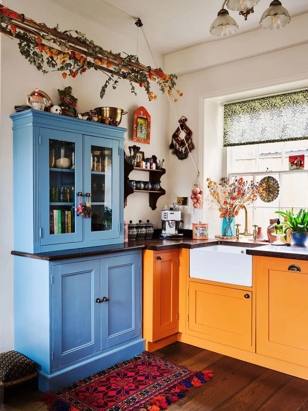 A blue glass-fronted kitchen dresser stands beside orange sink cabinets, a patterned blind, hanging decorative objects and a red patterned rug