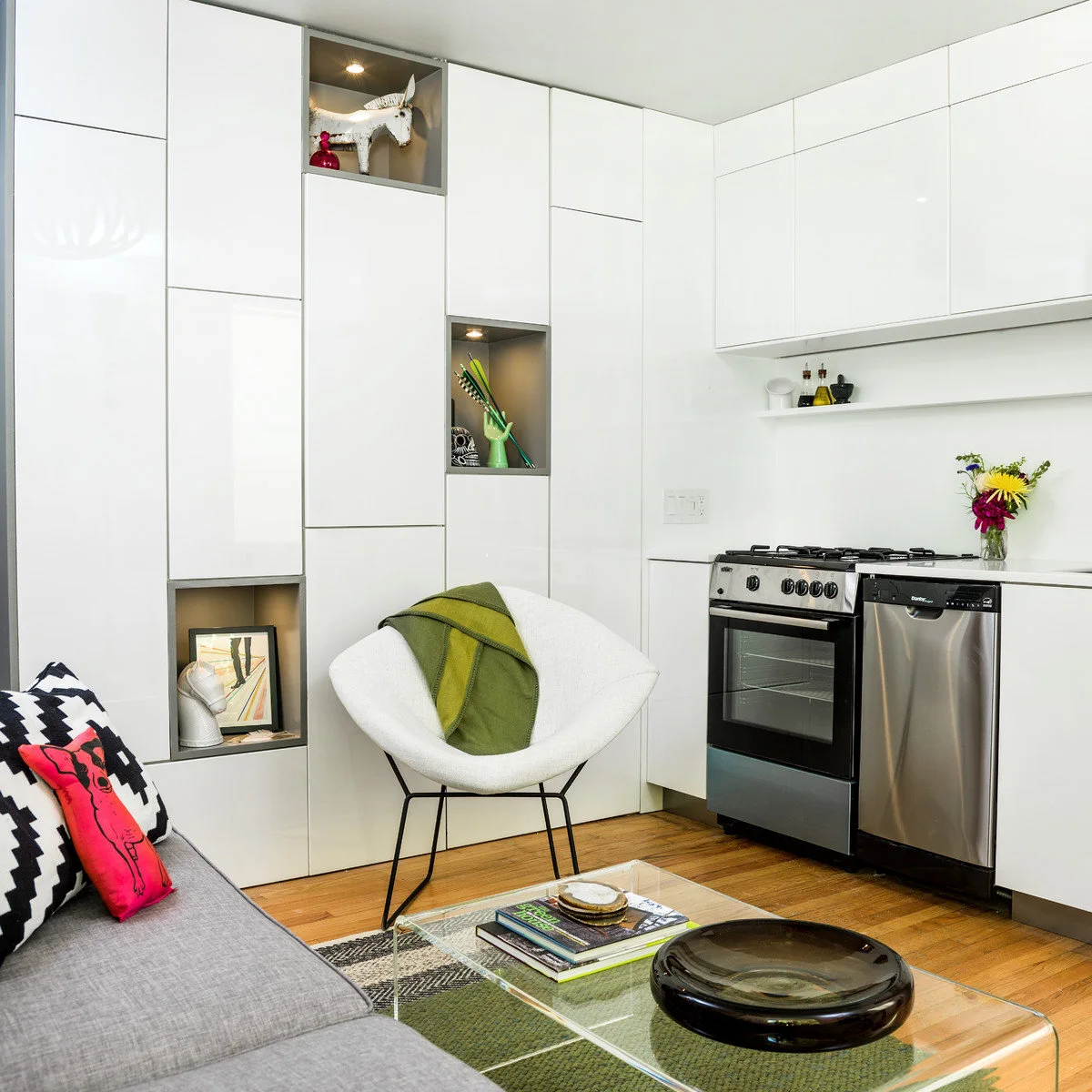 Small studio with white kitchen cabinets continuing into a tall storage wall beside a sofa and chair