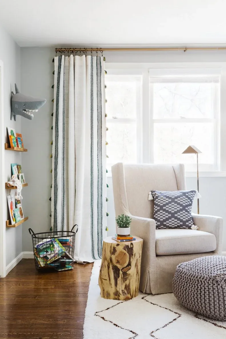 Pale adult armchair beside wood-toned cylindrical table, knitted-looking pouf, book basket, patterned curtains and pale rug.