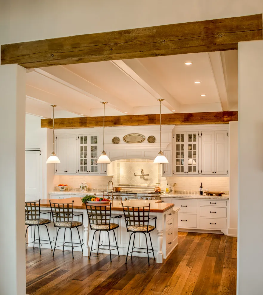 White cabinetry fills a kitchen below broad timber beams beside a small dining island