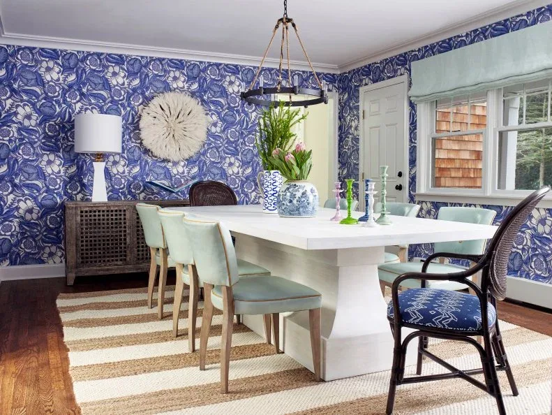 Dining room with blue patterned walls, white pedestal table, pale aqua chairs, dark woven end chairs and striped rug.