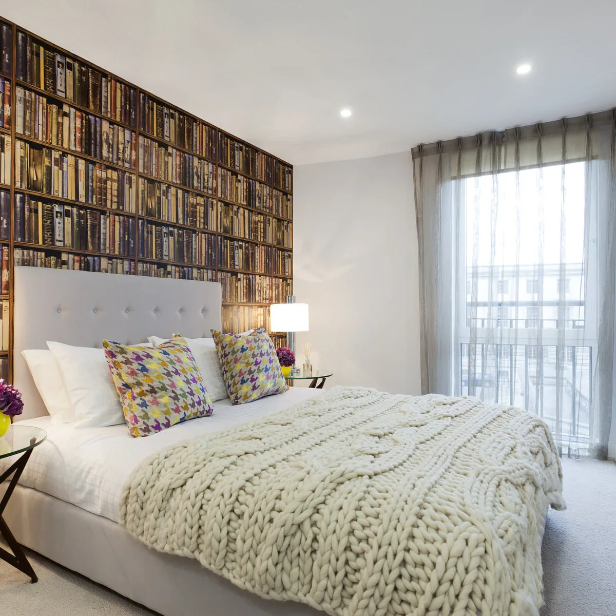 Bookshelf-print wallpaper behind a pale gray headboard with white bedding, a chunky throw and colorful cushions.