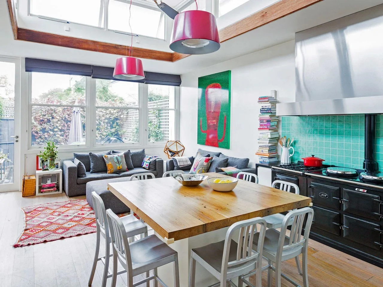 A timber dining table stands between a dark range and gray sofas beneath high windows and pink pendant lights.
