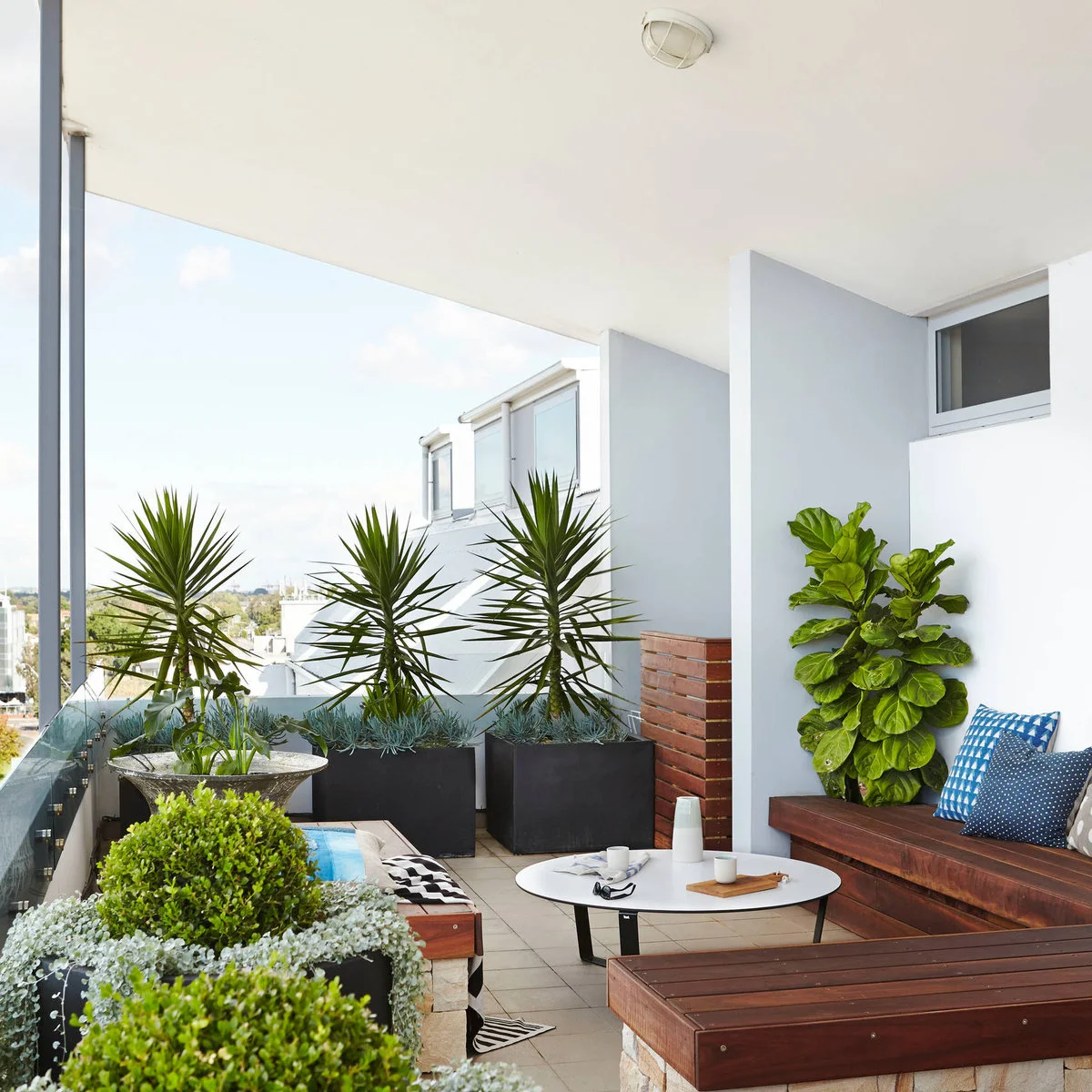 Apartment balcony with wood-colored benches, low oval table and several large planters