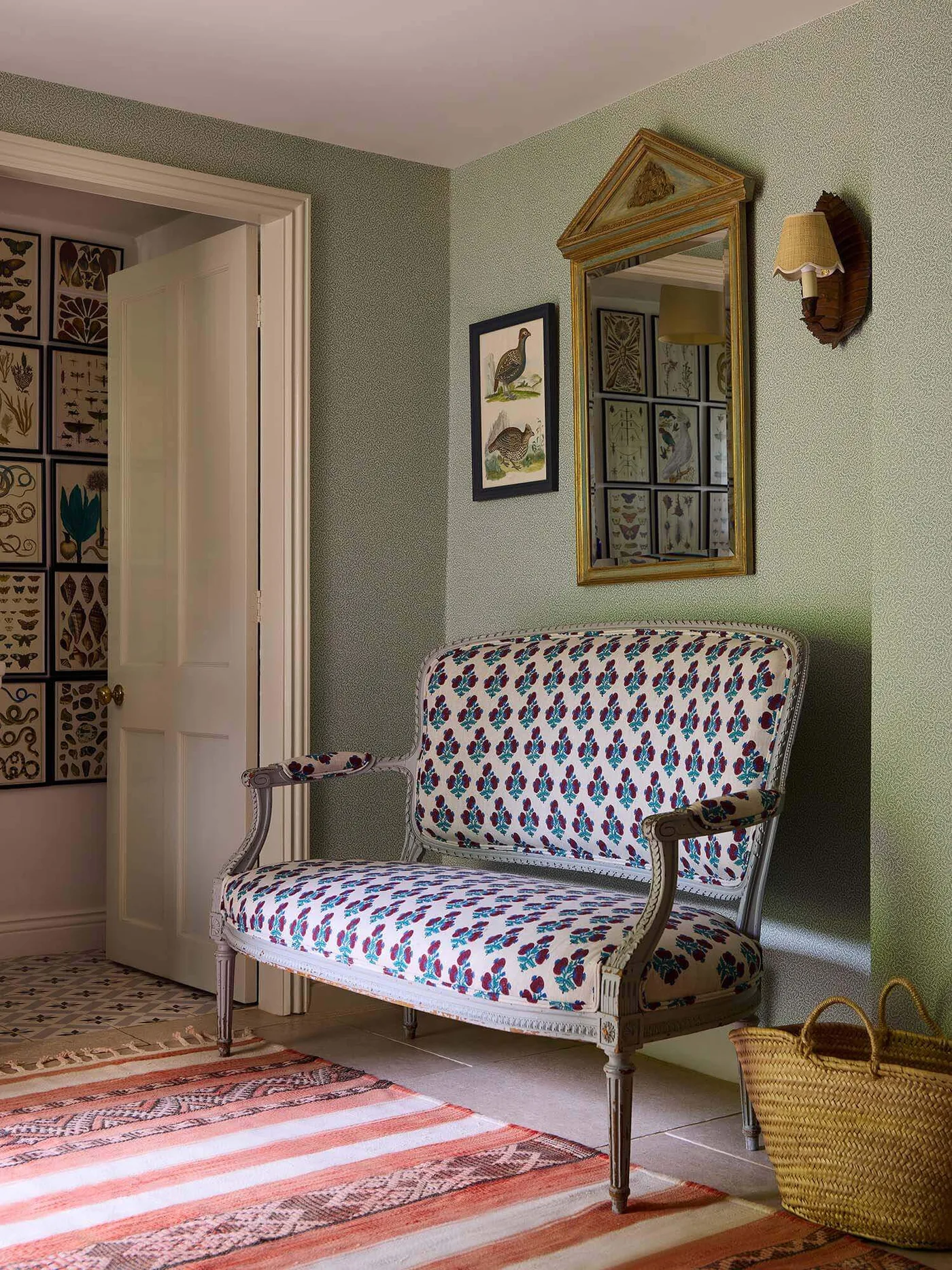 Hallway with pale green walls, patterned upholstered bench, tall shaped mirror, small wall light, striped runner and woven basket.