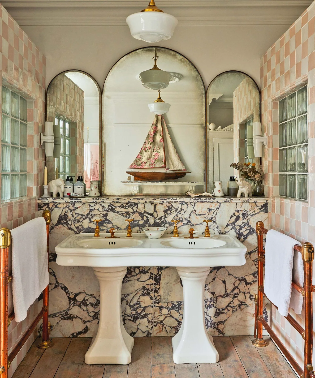 A double pedestal basin stands before strongly veined lower walls and pale pink checked tiles, beneath a three-part arched mirror and beside warm-metal towel rails.