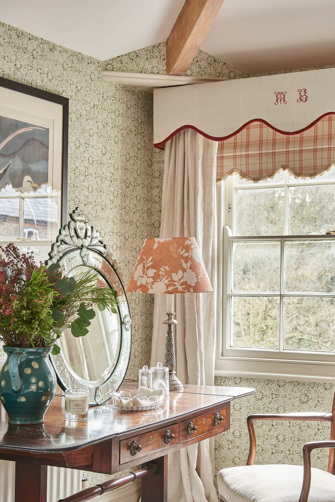 A dark timber dressing table with an ornate oval mirror stands beside a window and patterned lamp shade