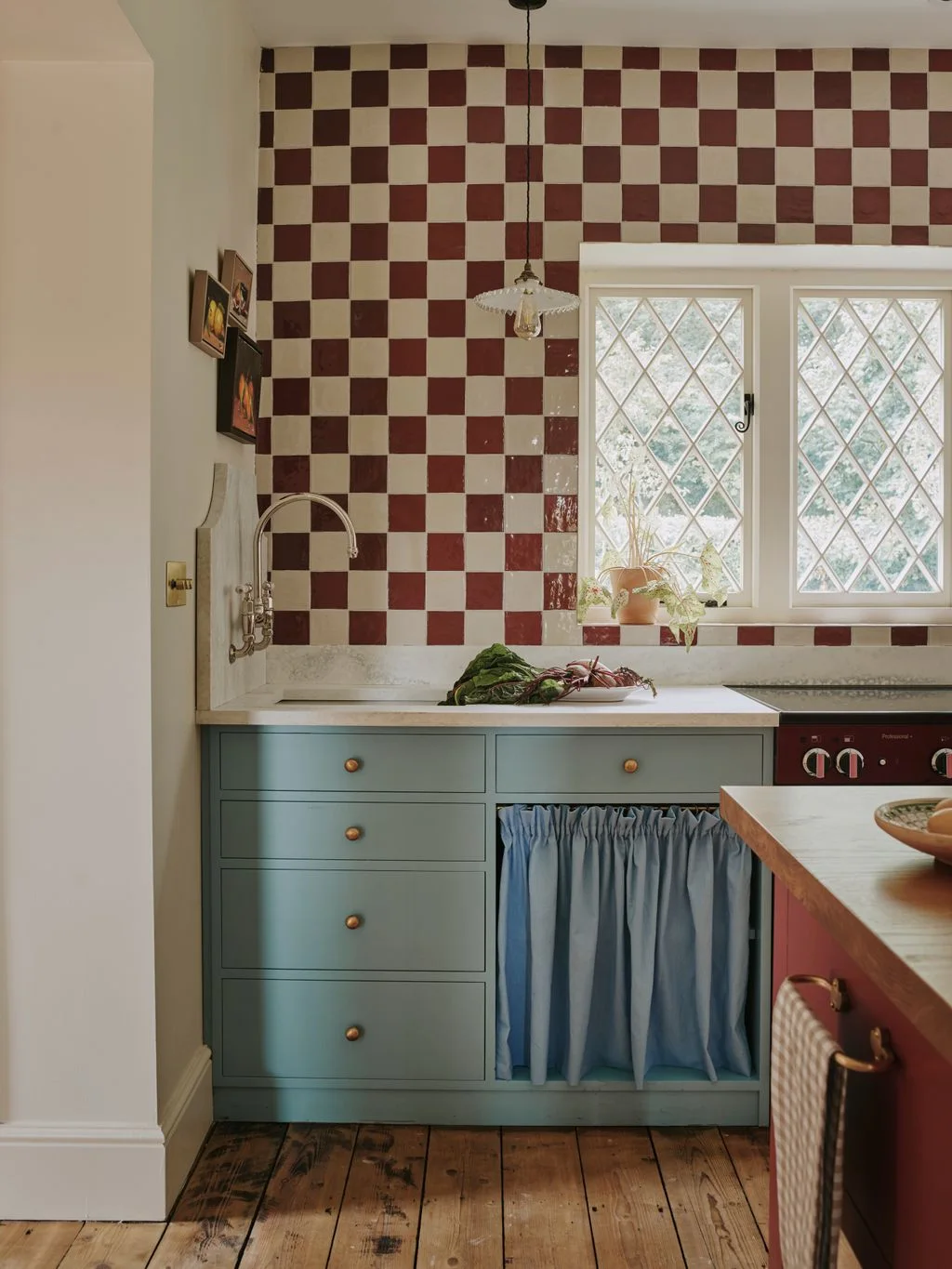 Blue-gray kitchen cabinets with a gathered skirt sit beneath red-and-cream checked wall tiles around a small leaded window.