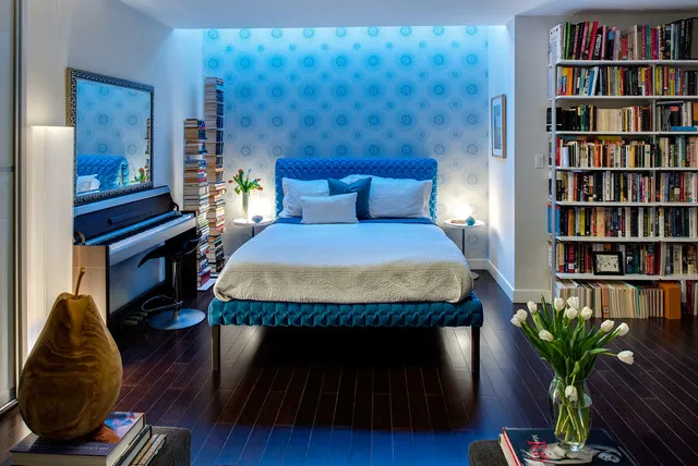 Small blue motifs cover the wall behind a bright blue bed near tall bookshelves.