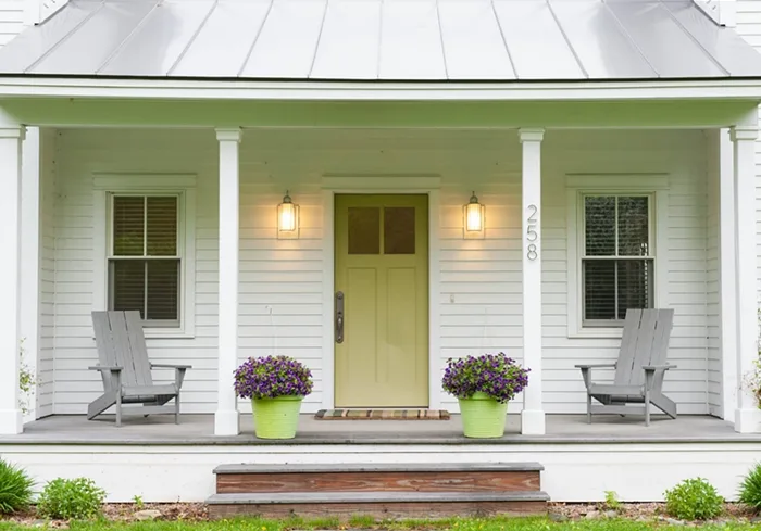 White farmhouse porch with yellow-green door, paired gray chairs and green flowerpots