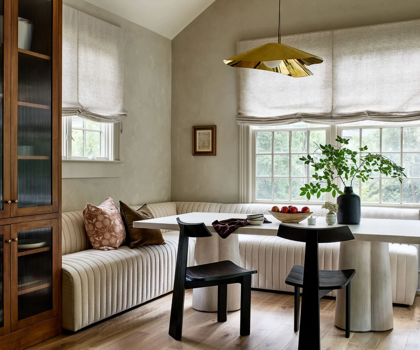 A pale upholstered corner banquette and white oval table sit beside dark chairs, wood cabinetry and soft window blinds.