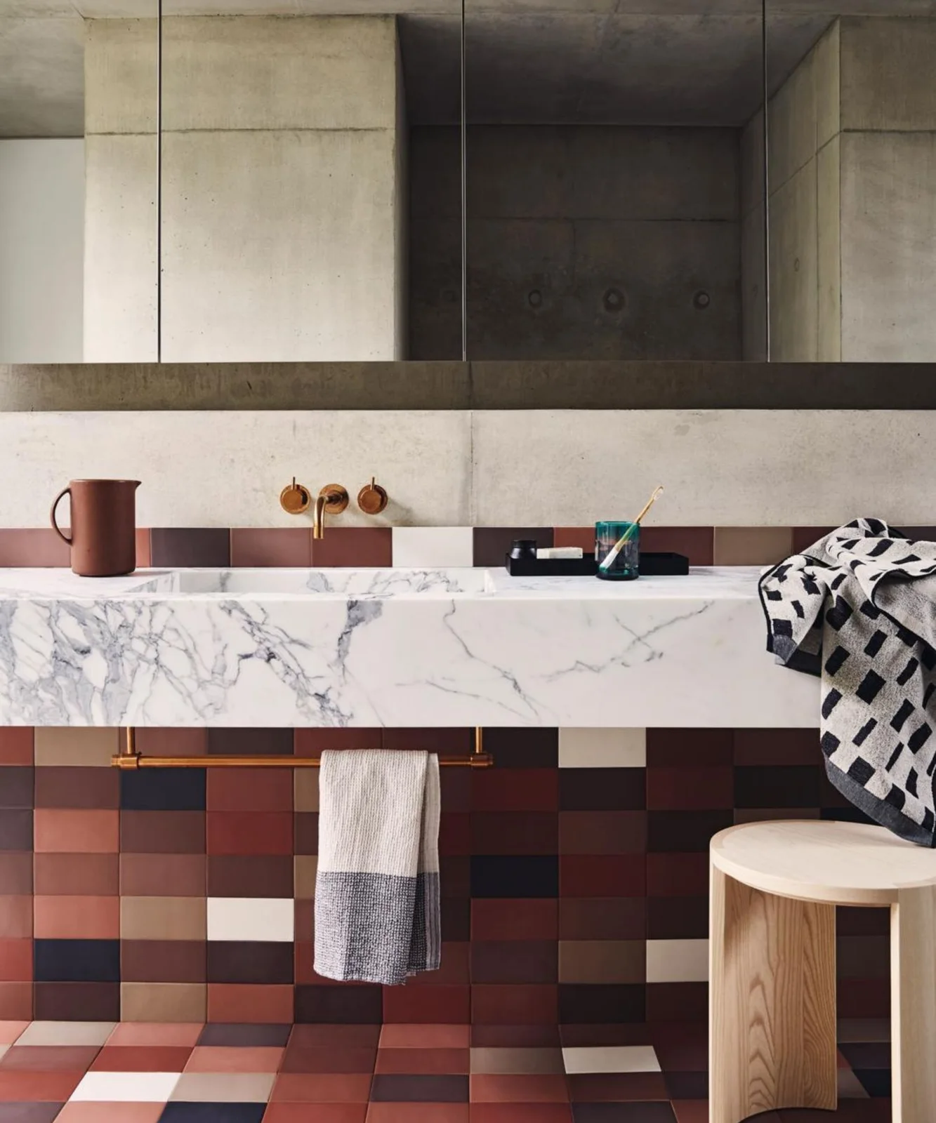 A pale veined floating bathroom basin sits above warm checker-patterned wall and floor tiles beneath a wide mirror beside a small wooden stool