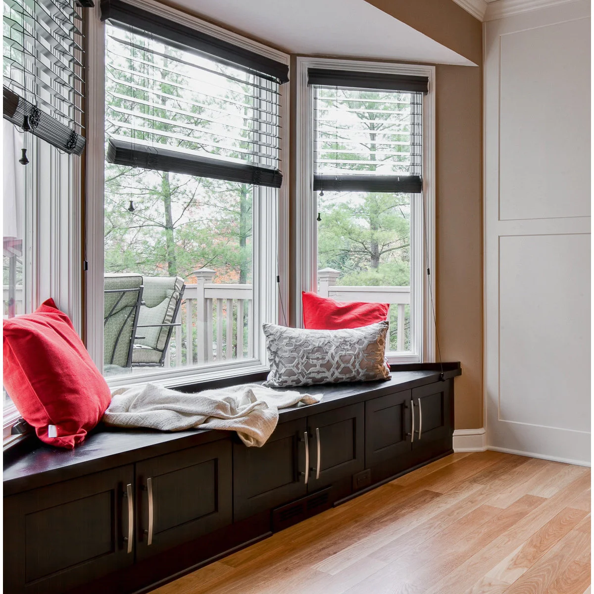 A dark storage bench beneath an angled bay window, with red cushions and black slatted blinds.
