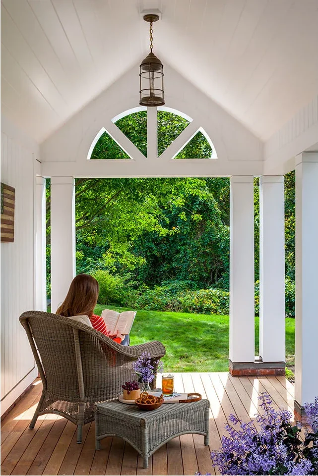 A woven chair and footstool sit beneath a white gabled porch ceiling with an arched opening, hanging lantern and a view of the garden.