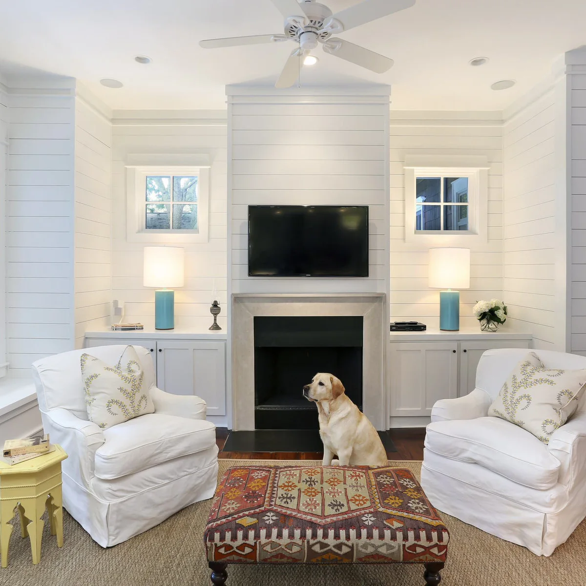 White paneled sitting area with two white chairs, a patterned ottoman and fireplace