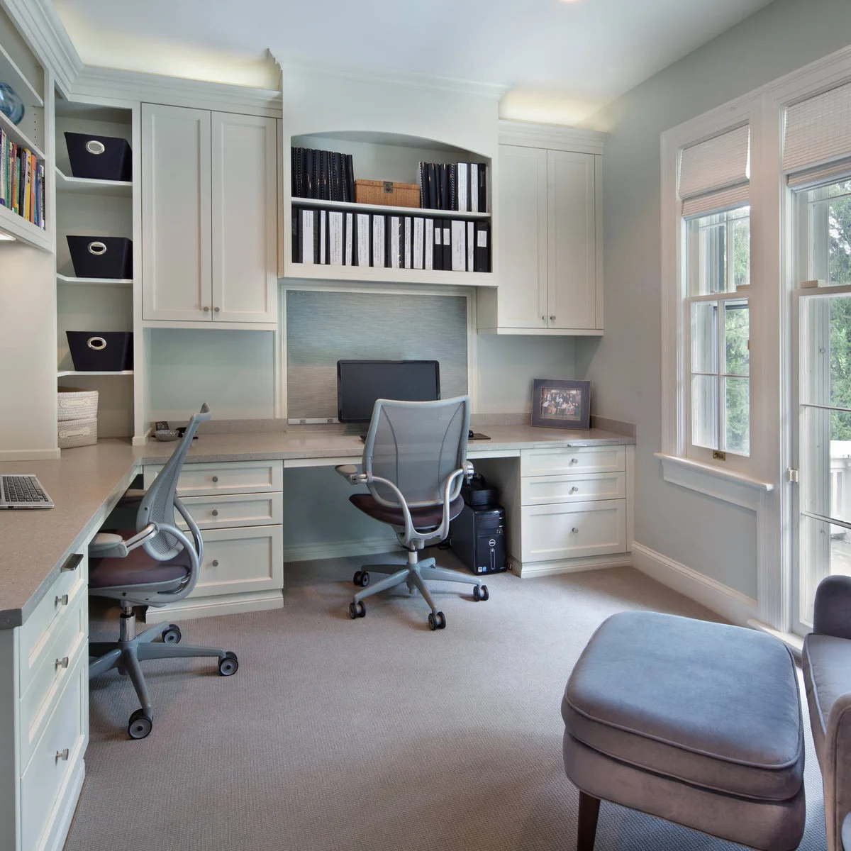 Fitted corner desks with white cabinetry, two adjustable chairs and tall windows beside a glazed door