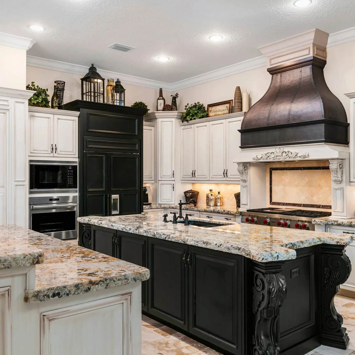 Black lanterns, vessels, greenery and a framed sign above stepped white and black kitchen cabinets beside a large dark hood.