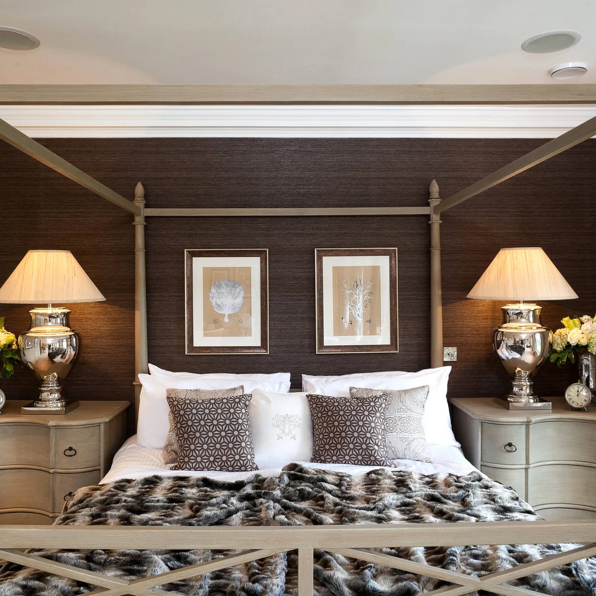 Dark brown textured wall behind a pale four-poster bed with light bedside chests and reflective lamps.