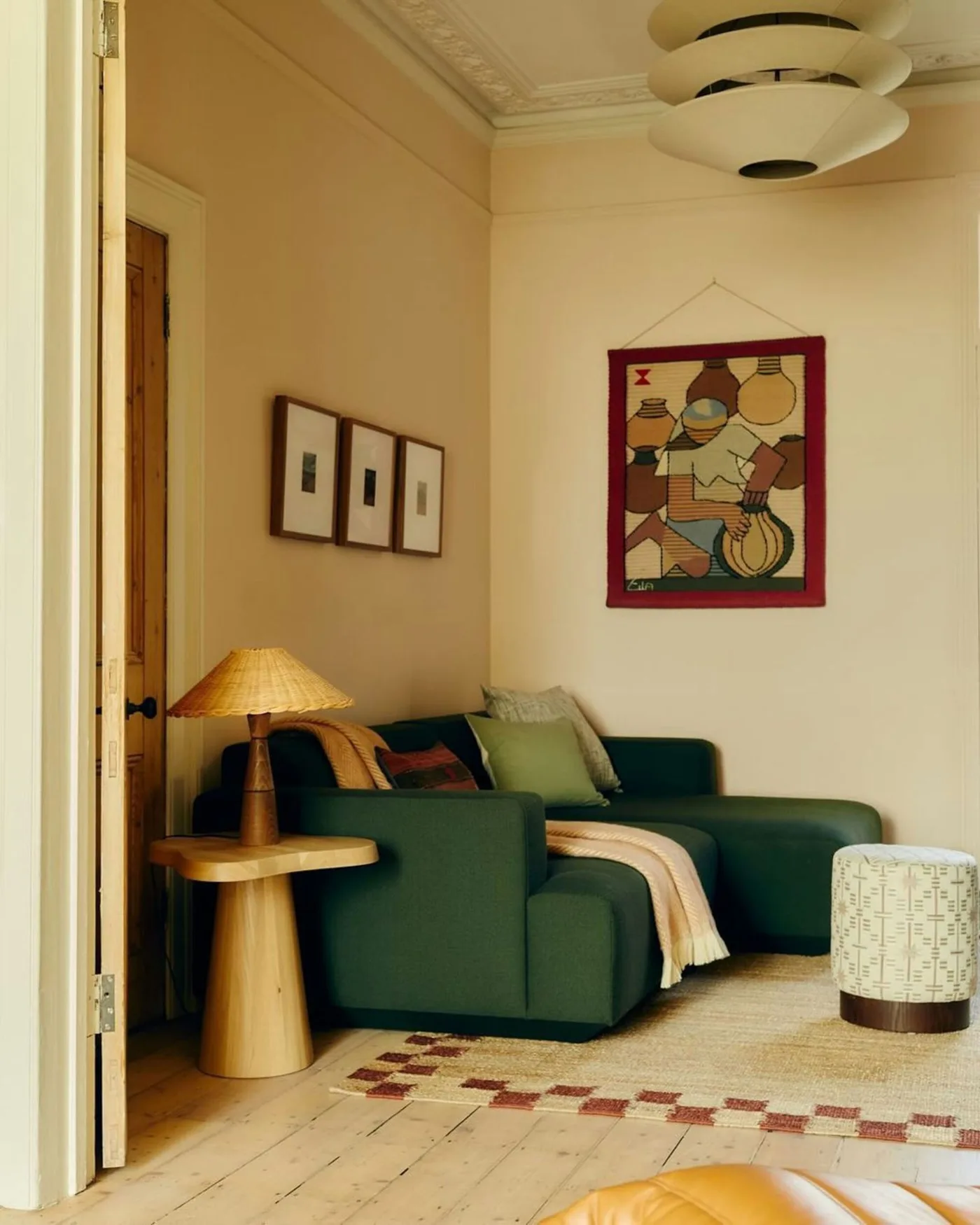 Deep green sofa in a warm pale living room with a wood side table, shaded lamp and checked rug border