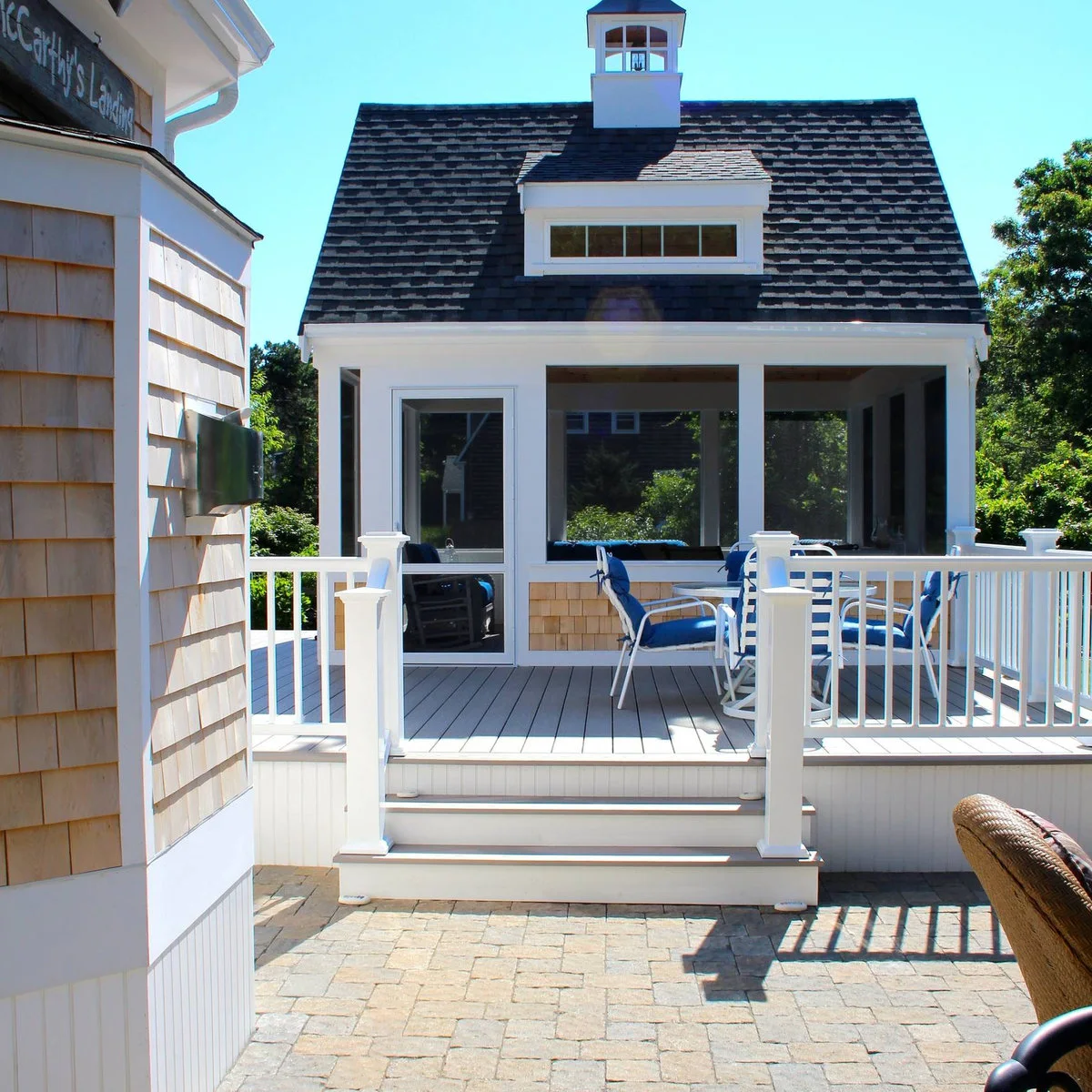 A screened porch with a steep roof and small cupola opens onto a white-railed deck with a round table and blue-cushioned chairs.