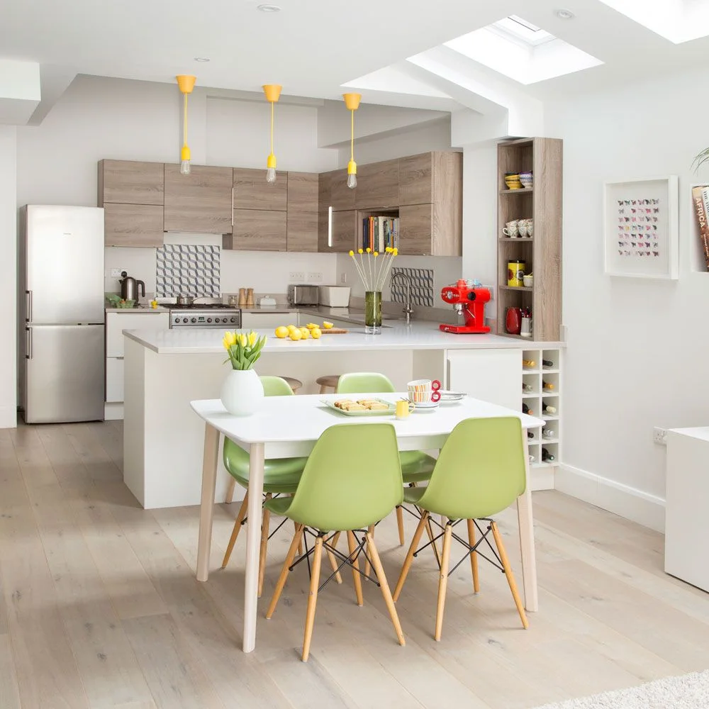 A white dining table with pale green chairs stands beside a white peninsula and wood-toned kitchen cabinets under skylights