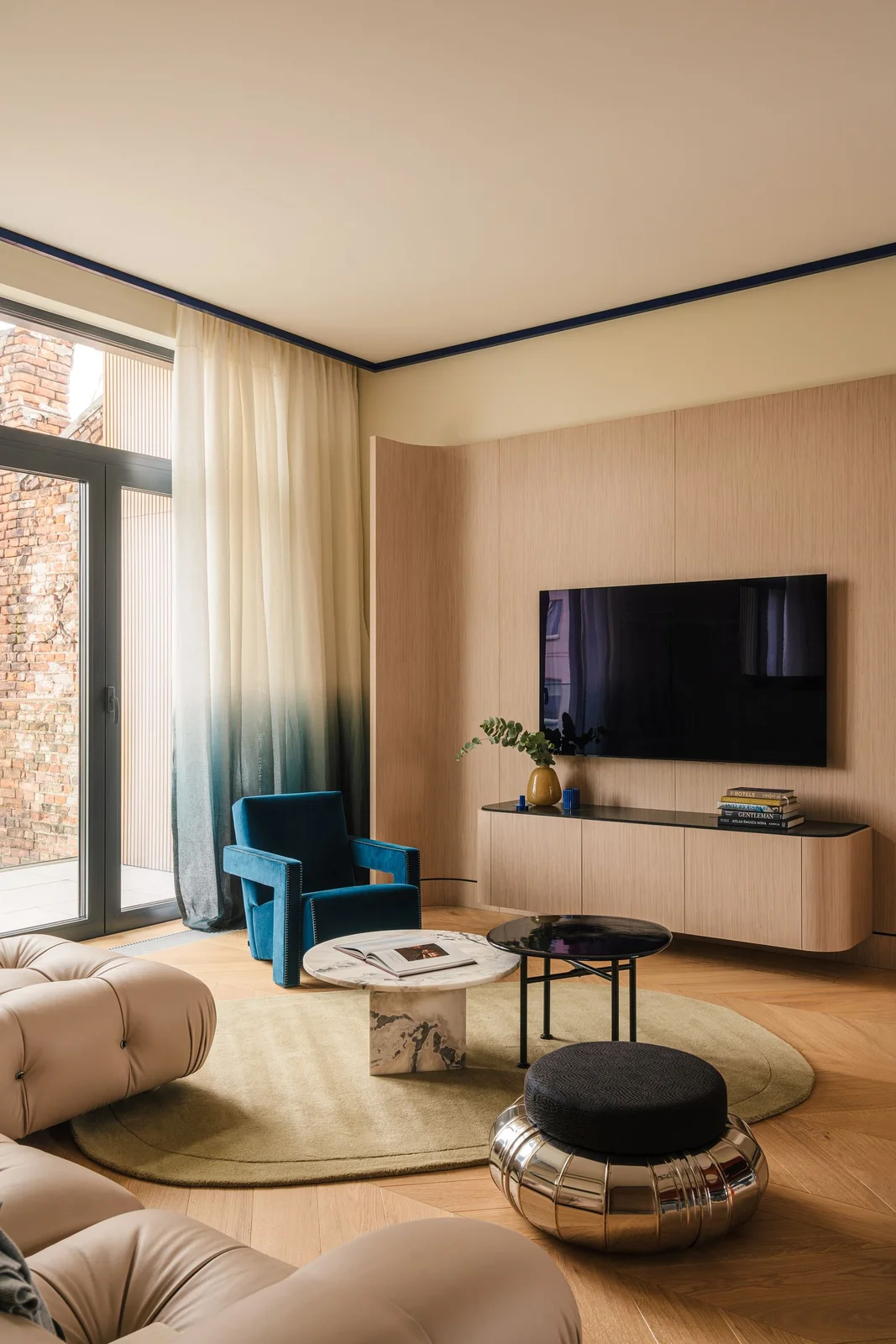 A television hangs on a curved wooden wall above a matching suspended cabinet beside a blue armchair and pale curtains