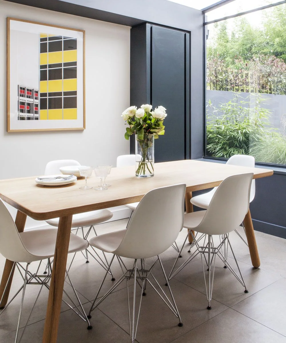 A pale rectangular wood table and white chairs with slim metal bases sit beside a garden window, dark storage doors and geometric artwork.