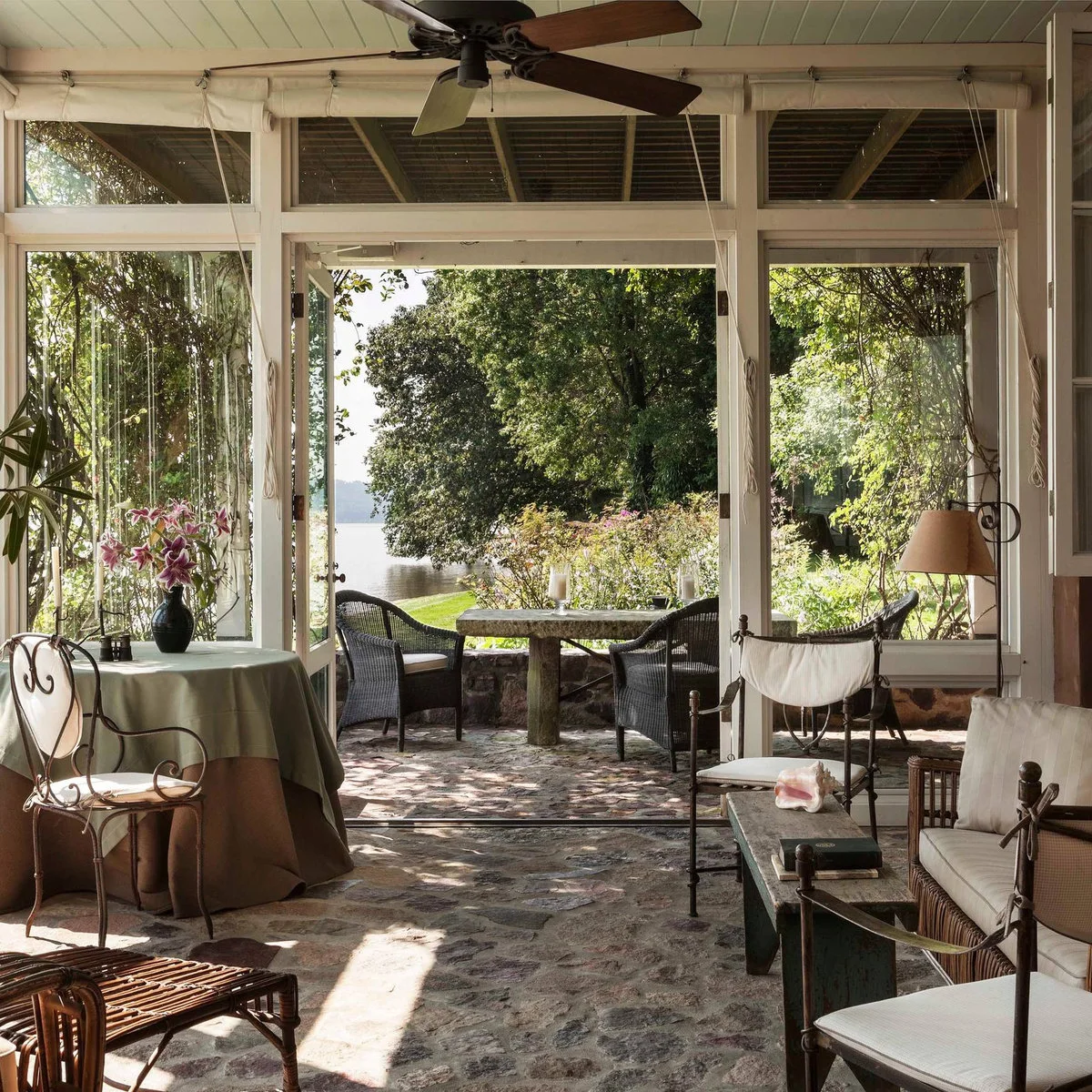 A porch with irregular stone flooring, light-cushioned chairs and a broad open doorway leading to outdoor seating.
