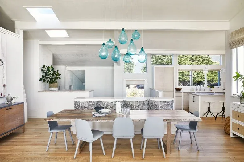 Open dining area with long wooden table, gray and white chairs, patterned bench and clustered blue glass pendants.