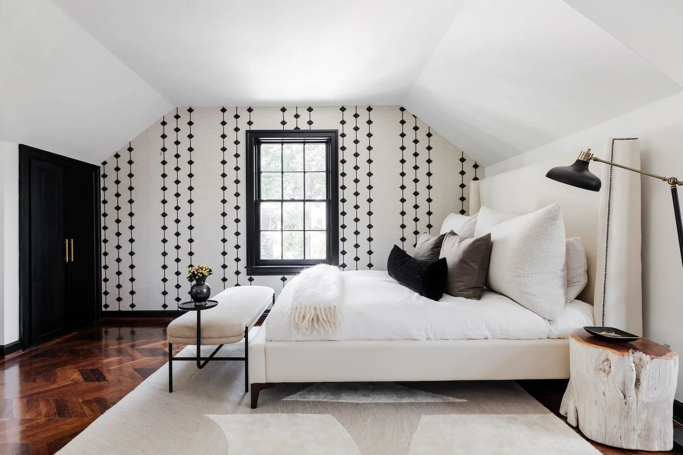 A white bedroom has a black-framed window, vertical black motif wallpaper, a pale upholstered bed and a warm wood floor beside a dark wardrobe