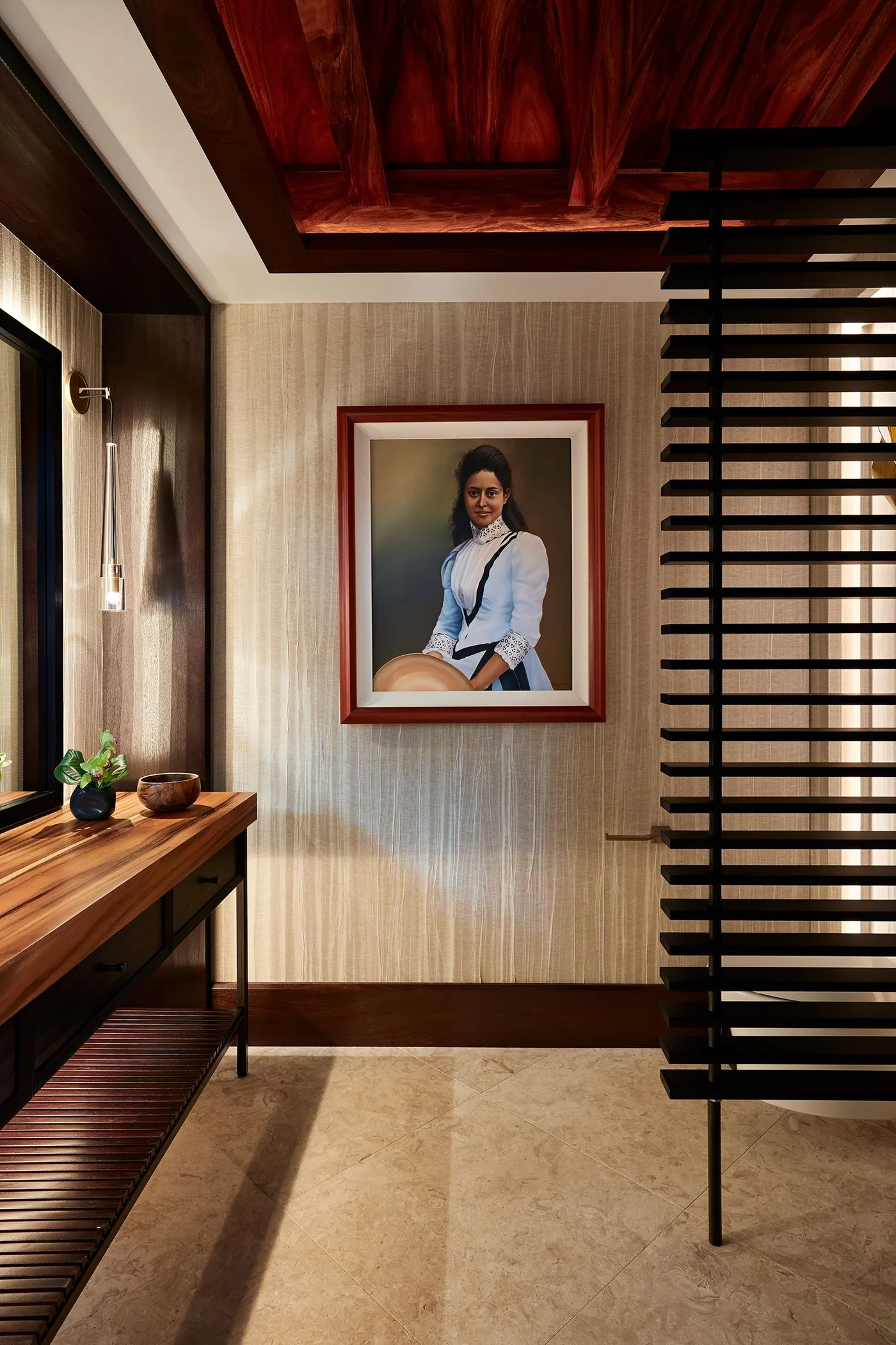 Powder room with textured pale walls, framed portrait, wooden counter and lower shelf, dark slatted screen, warm wooden ceiling and small plant.